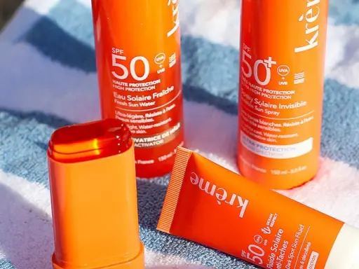 Trois produits solaires orange vif, dont un stick, sur une serviette rayée. Idéal pour savoir quelle crème solaire choisir pour une haute protection SPF 50.
