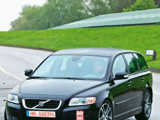 Czarne Volvo V50 2.0D na mokrej drodze, za kierownicą siedzi mężczyzna. W tle zielone wzgórza i budynek.
