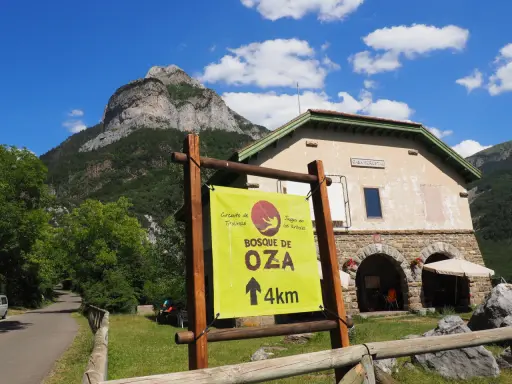 Cartel indica "Bosque de Oza 4km" en un día soleado en el valle de hecho, con una casa forestal y montañas al fondo.