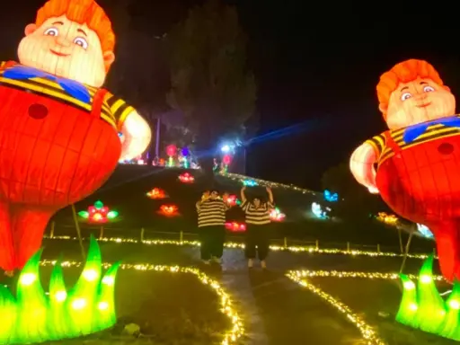 Gigantesche lanterne di bambini paffuti illuminano un sentiero a Wonderland Rimini, con due persone che salutano tra fiori luminosi.