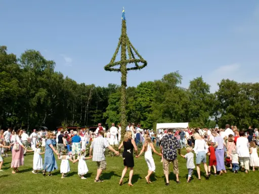 Människor dansar runt en midsommarstång i en grönskande park. Många barn med blomsterkransar i håret deltar i firandet.