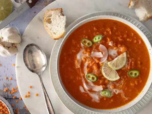 Eine cremige rote Linsensuppe mit Kokosmilch, garniert mit Limettenspalten und Jalapeños. Dazu gibt es knuspriges Brot.
