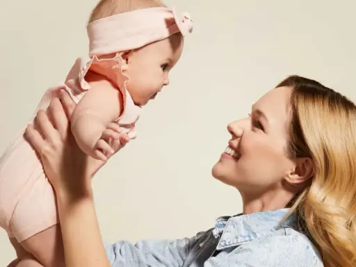 Mama podnosi dziecko w różowym body i opasce. Idealny przykład, jak ubierać noworodka latem – lekko i wygodnie.