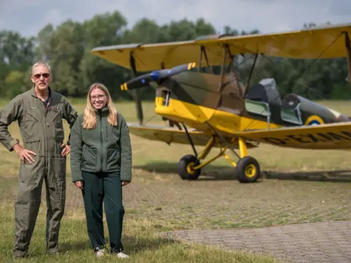 Mann in Fliegerkluft und junge Frau stehen neben gelbem Doppeldeckerflugzeug. Das Flugzeug erinnert an einen Tiger Moth.