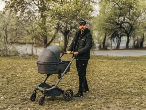 Mężczyzna w czapce z daszkiem i kurtce pcha nowoczesny wózek dla dziecka po trawie.