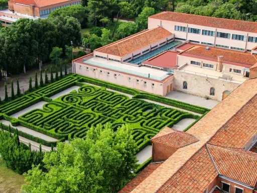 Un giardino con un intricato labirinto borges, circondato da edifici con tetti rossi e alberi verdi.