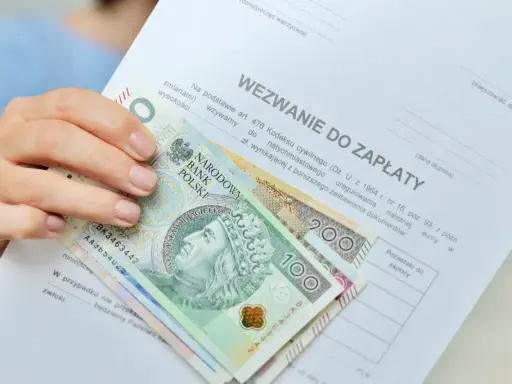 Dłoń trzyma wezwanie do zapłaty i polskie banknoty 100 i 200 zł. Dokument zawiera informację o zawiadomieniu o uchyleniu zajęcia.