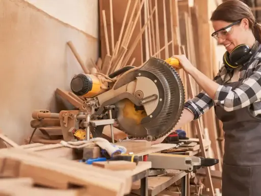 Artisane souriante utilisant une scie à onglet. Elle porte des lunettes et un tablier, des éléments essentiels pour comment protéger une création artisanale.
