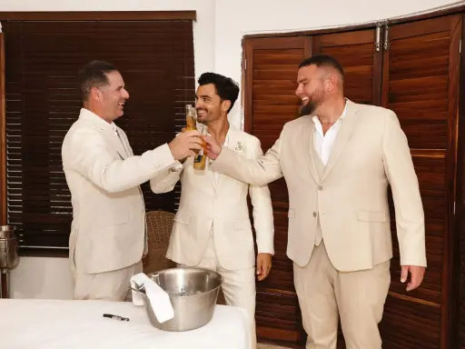 Tres hombres en trajes claros brindan con cerveza. El novio, en el centro, sonríe. Un momento de alegría antes del discurso de boda hermano.