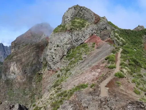 Steiler Bergpfad auf Madeira, ideal für die Packliste. Felsige Gipfel, grüne Vegetation und ein Weg, der sich durch die zerklüftete Landschaft schlängelt.
