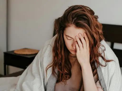 Kvinna med rött hår i sängen, täckt av filt, håller handen för ansiktet. Hon ser ut att ha feber, ett kolera symptom.