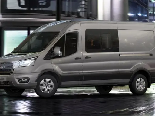 Camión N1 gris Ford Transit circulando por una calle urbana mojada de noche, con luces de edificios y tráfico difuminadas al fondo.