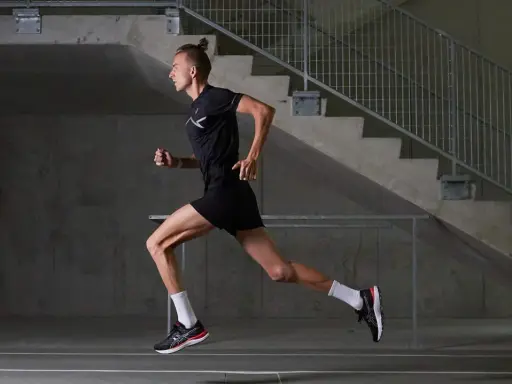 Mężczyzna w czarnym stroju sportowym biegnie, czerpiąc z biegania w tlenie. Dynamiczna sylwetka na tle betonowych schodów.
