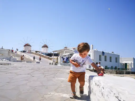 Un bambino gioca con una macchinina rossa a Mykonos, con i mulini a vento sullo sfondo. Vacanze a Mykonos con bambini.