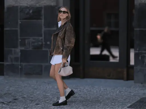 Stylish woman in a white dress, brown leather jacket, and black loafers with white socks walks confidently.