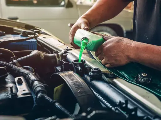 Mecánico rellenando refrigerante verde en el radiador de un coche, solucionando una posible fuga de gas aire acondicionado.