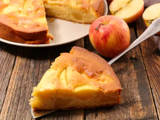 Una fetta di torta di mele senza burro, dorata e soffice, con mele a vista. Accanto, una mela rossa e gialla e altre fette.