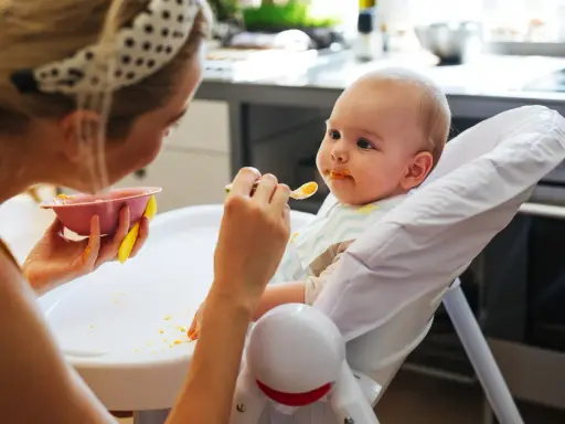 Mama karmi 7 miesięczne dziecko puree z marchewki. Maluch ma pomarańczowe ślady na buzi i śliniaczku.