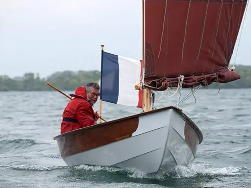 Un homme en ciré rouge navigue sur son bateau, fier du résultat de son travail pour construire son bateau. Le drapeau français flotte fièrement.