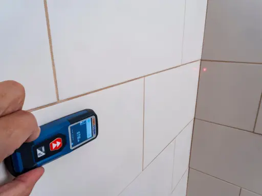 Mano sujetando un aparato para medir con láser rojo apuntando a la esquina de una pared con baldosas blancas.