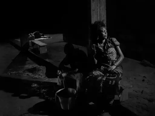 A woman in a patterned dress sits in shadow, eyes closed, while a man works with a bucket in the foreground. Alex Majoli captures a moment of quiet resilience.
