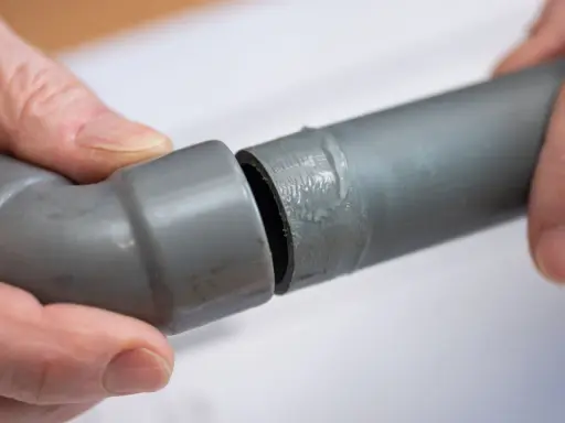 Mains de personne assemblant deux tuyaux gris en **pvc renforcé**, l'un avec un coude, prêts pour une installation solide.