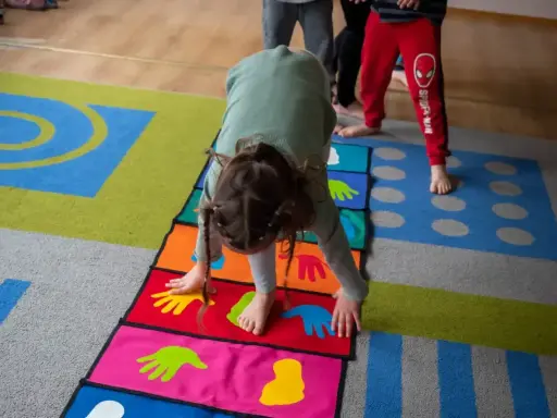 Dziecko wykonuje ćwiczenia na kolorowej macie z symbolami dłoni i stóp. Poduszka sensoryczna wspiera rozwój motoryczny.