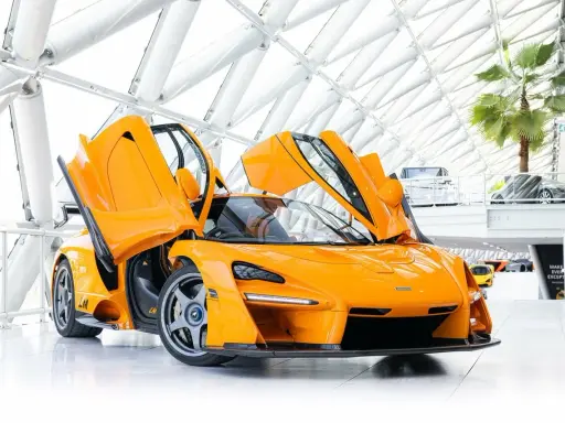 Ein leuchtend oranger McLaren Senna LM mit geöffneten Flügeltüren steht in einem modernen Gebäude mit Glasdach.