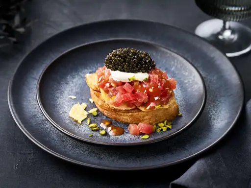 Kaviar Rezepte Vorspeise: Thunfisch-Tatar auf geröstetem Brot, gekrönt mit Kaviar und einem Klecks Crème fraîche. Pistazien und Chips garnieren den Teller.