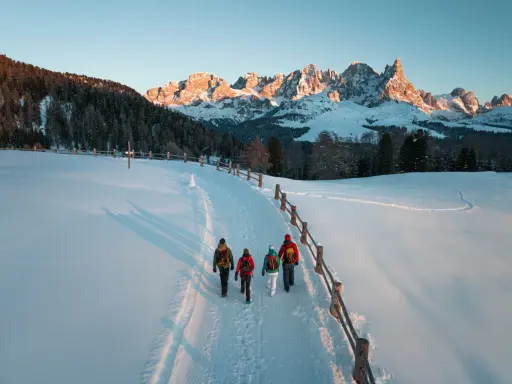 Quattro escursionisti camminano su un sentiero innevato al Passo di Lavazè, con le maestose vette dolomitiche illuminate dal sole al tramonto.