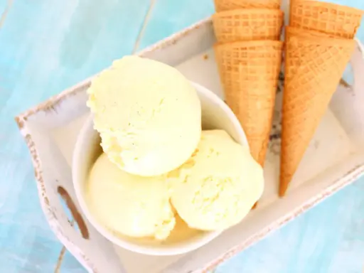 Tres bolas de cremoso helado de vainilla en un cuenco blanco, junto a conos de galleta sobre una bandeja.