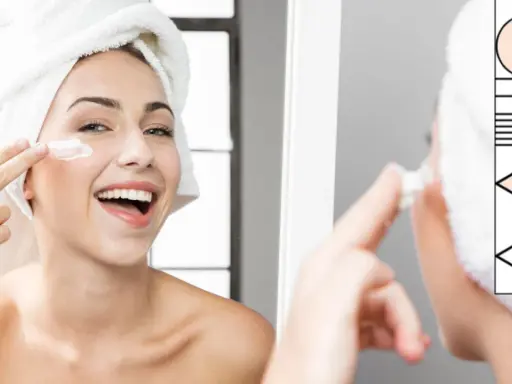 Donna sorridente con asciugamano in testa applica crema sul viso, parte della sua routine skincare.