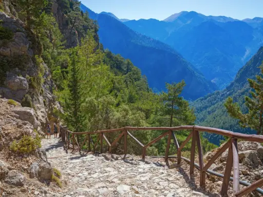 Wanderweg in einer atemberaubenden **Schlucht auf Kreta**. Steile Felswände, grüne Bäume und ein tiefer Abgrund, gesichert durch einen hölzernen Zaun.