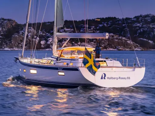 Eine Hallberg-Rassy 69 gleitet bei Dämmerung über das Wasser, die schwedische Flagge weht im Wind.