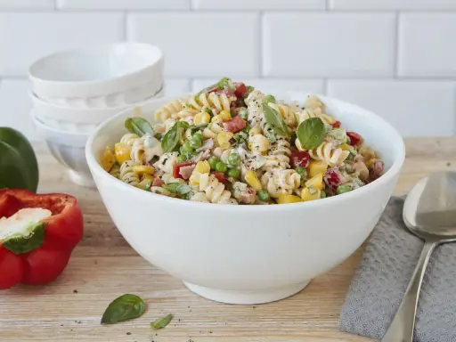 Ein bunter Nudelsalat ohne Mayonnaise mit Spiralnudeln, Erbsen, Mais, Paprika und frischem Basilikum in einer weißen Schüssel.