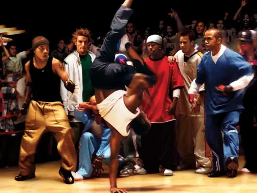 Energetyczna scena z filmów o tańcu. Breakdancer wykonuje akrobację, otoczony przez widzów i innych tancerzy.