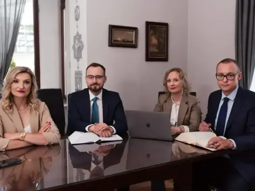Cztery osoby w strojach biznesowych siedzą przy stole, przed nimi dokumenty i laptop.