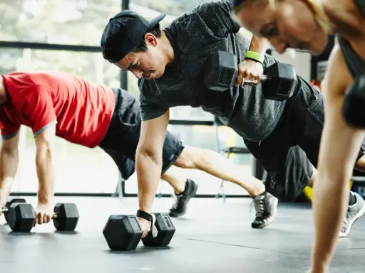 Drei Personen machen ein intensives Crossfit Training mit Kurzhanteln. Sie sind in einer Plank-Position und ziehen abwechselnd eine Hantel hoch.