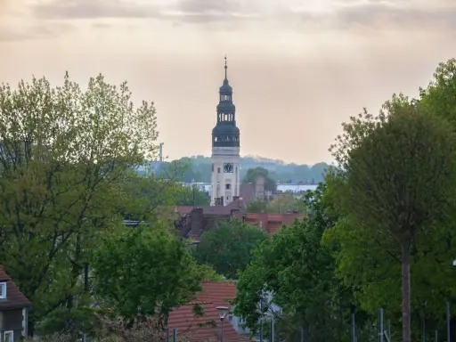 Wieża zegarowa góruje nad zieloną górą, skąd rozciąga się widok na rynek i dachy domów.