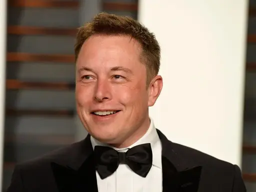 Uśmiechnięty Elon Musk w smokingu z muszką. Wiek nie jest widoczny, ale jego twarz wyraża pewność siebie.