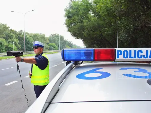 Policjant z radarem na drodze, obok radiowozu z napisem POLICJA.