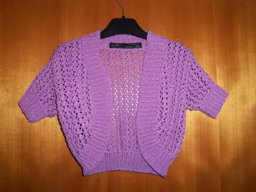 A purple crocheted bolero with short sleeves and an openwork pattern hangs on a hanger against a wooden background.
