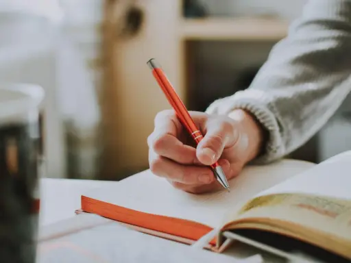 Main tenant une plume rouge, une main écrit dans un carnet. Un synonyme de participer pourrait être collaborer, comme dans cette scène d'étude.