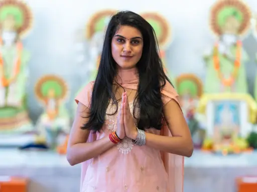En ung kvinna i traditionella kläder med händerna i bön framför hinduiska statyer, en bild som väcker tankar om mantra hinduism.