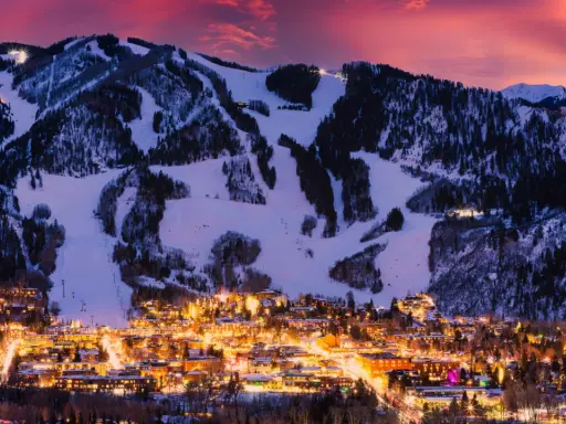 Glitzerndes Aspen, Colorado, im Abendrot. Schneebedeckte Berge mit Skipisten über einer Stadt, die in warmem Licht erstrahlt.