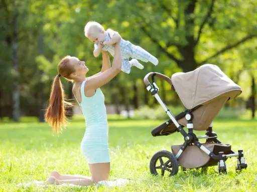Uśmiechnięta mama podrzuca dziecko w powietrze na trawie obok wózka.