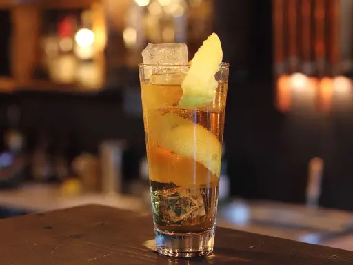 Ein Longdrink mit Eis, Zitronenzeste und einer Orangenscheibe. Die Zitrone windet sich wie ein Pferdehals im Glas.