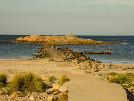En stenpir leder ut till en klippö. En person går på piren, en annan badar. Perfekt för aktiviteter i Halmstad.