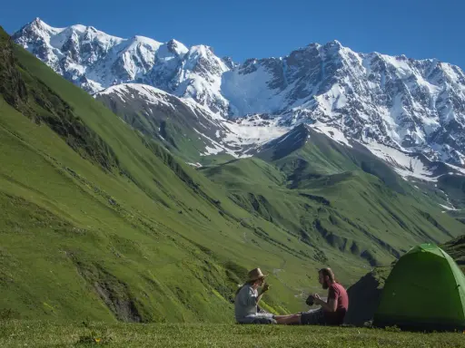 Para odpoczywa po trekkingu w Gruzji, siedząc obok zielonego namiotu na tle majestatycznych, ośnieżonych gór.