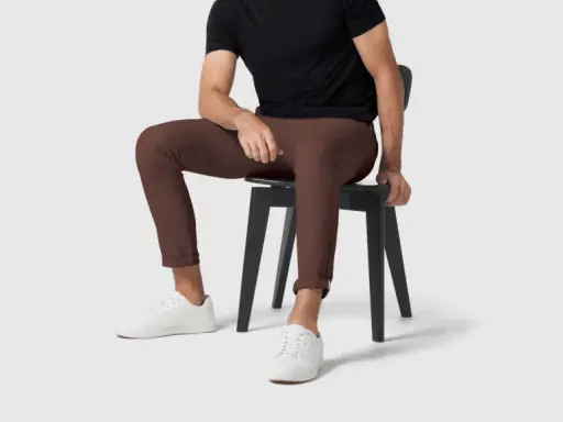 A man in a black shirt and brown pants sits on a black chair, wearing white sneakers.